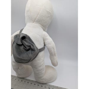 May include: A large, white plush toy in a standing pose, with a gray backpack. The toy has a rounded head and body, with a soft, textured surface. The backpack is attached with gray straps and has a curved flap.