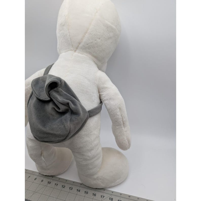 May include: A large, white plush toy in a standing pose, with a gray backpack. The toy has a rounded head and body, with a soft, textured surface. The backpack is attached with gray straps and has a curved flap.
