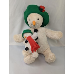 May include: A plush snowman toy with a green hat and a red flower. The snowman has a carrot nose and black button eyes. A smaller snowman with a red scarf is held by the larger snowman. The toy is cream-colored.
