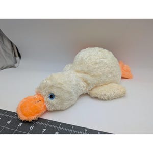 May include: A cream-colored plush duck toy with a bright orange bill and feet, featuring blue embroidered eyes. The toy is positioned next to a black ruler, showing a length of approximately 11 inches from the bill to the tail end.