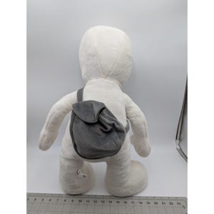 May include: A large, white plush figure with a gray backpack slung over its shoulder. The figure has a rounded head, arms, and legs. The gray backpack is small and has a flap closure. The figure is standing on a white surface.