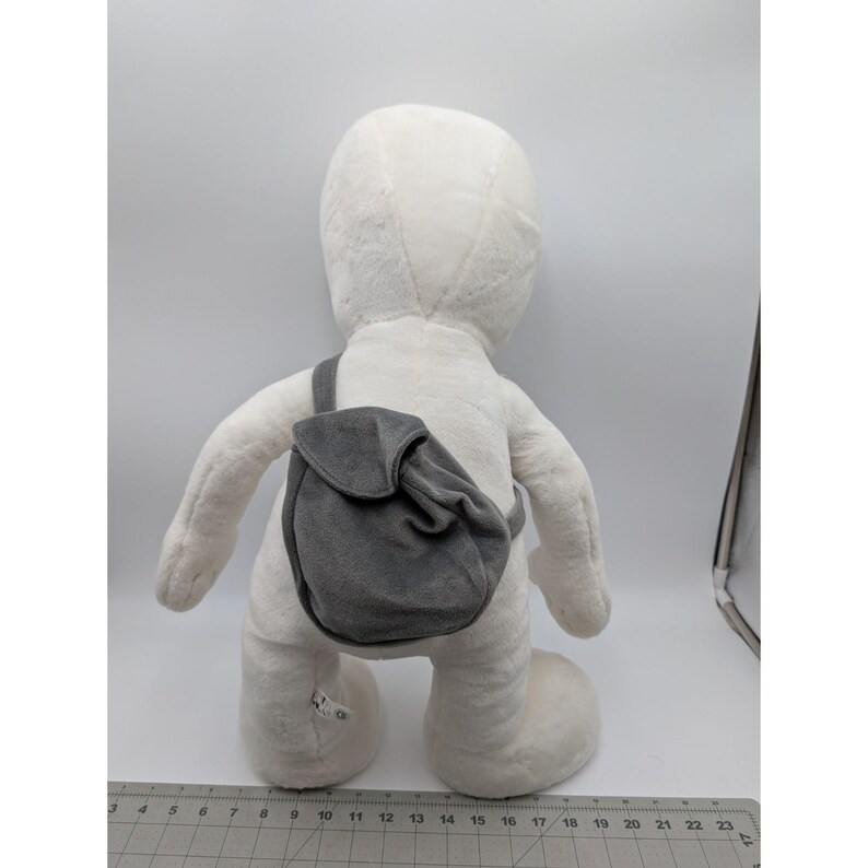 May include: A large, white plush figure with a gray backpack slung over its shoulder. The figure has a rounded head, arms, and legs. The gray backpack is small and has a flap closure. The figure is standing on a white surface.