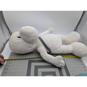 May include: A large, white plush toy in the shape of a cartoon character, lying on its side. The toy has large ears, a gray strap, and a smiling face. The toy measures approximately 15 inches in length.