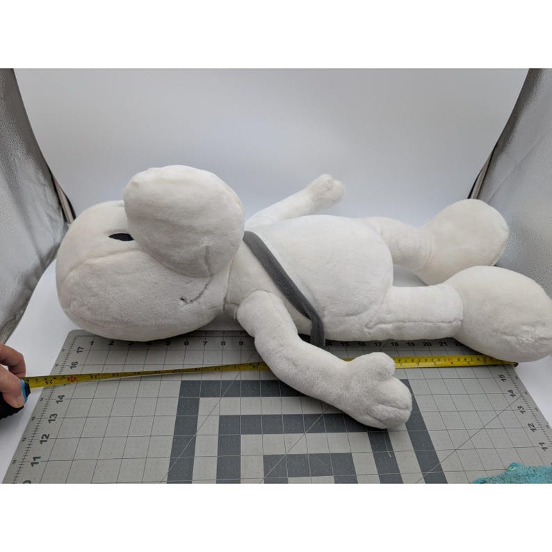 May include: A large, white plush toy in the shape of a cartoon character, lying on its side. The toy has large ears, a gray strap, and a smiling face. The toy measures approximately 15 inches in length.