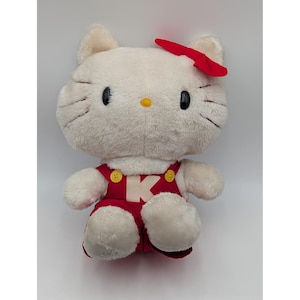 May include: A plush, white cat-like character with a red bow, black eyes, and a yellow nose. It wears a red overall dress with yellow buttons and the letter "K" on the front. The toy is soft and cuddly, perfect for children.
