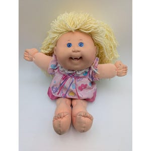 May include: A vintage Cabbage Patch Kid doll with blonde yarn hair, blue eyes, and a smiling expression. The doll wears a pink dress with a floral pattern and a white lace collar. Its arms and legs are outstretched.