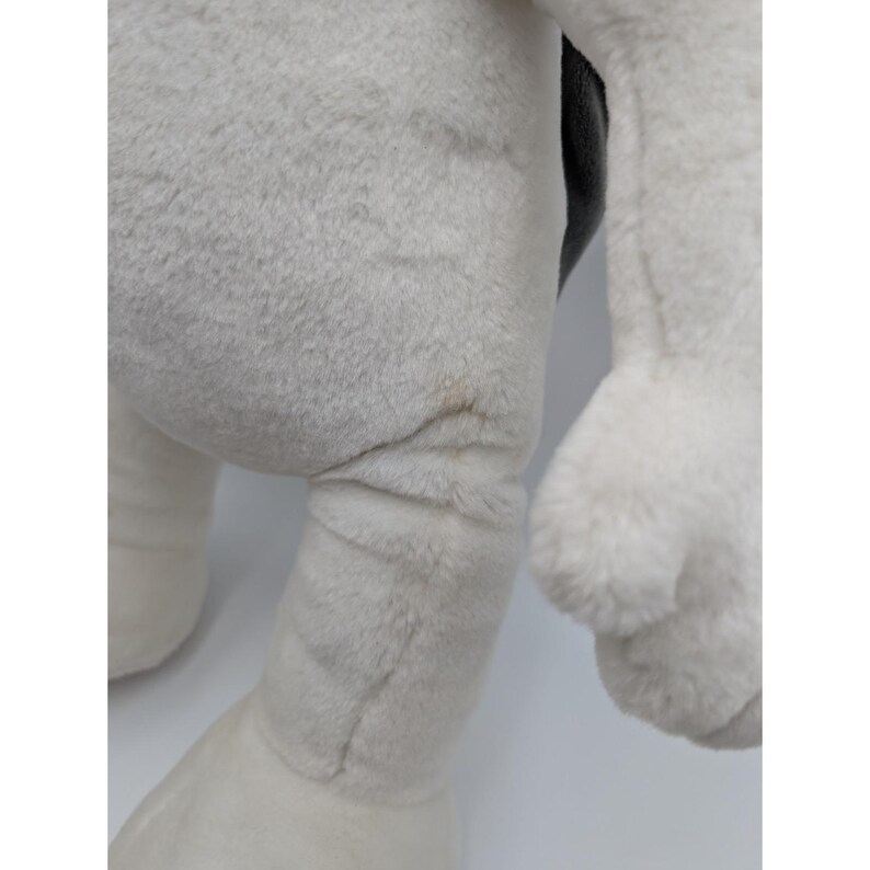May include: Close-up of a plush toy's leg and arm. The toy is primarily white with a gray area and a black section. The plush material appears soft and fluffy. There is a small brown stain on the leg.