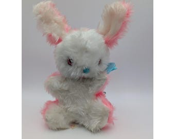 May include: A vintage stuffed bunny rabbit with white fur and pink accents on the ears, paws, and around the body. It has red eyes, a blue nose, and a small blue bow. The plush toy is sitting upright.