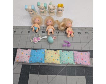 May include: A collection of vintage miniature toys, including small dolls with blonde hair and animal figurines. Accessories include a guitar, a pacifier, and a series of small pillows. The items are arranged on a gray cutting mat.