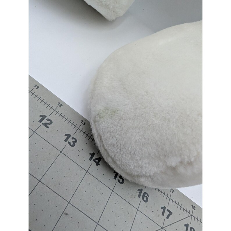 May include: Close-up of a white, fluffy, round object. The object has a soft, plush texture. A gray ruler with black markings is partially visible, showing measurements in inches.