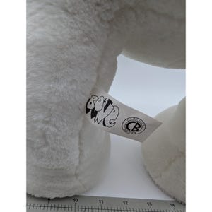 May include: Close-up of a white plush toy with a tag that reads "BONE" and "CARTOON BOOKS" in black text. The toy has a soft, fluffy texture. The tag is white with black text and a circular logo.