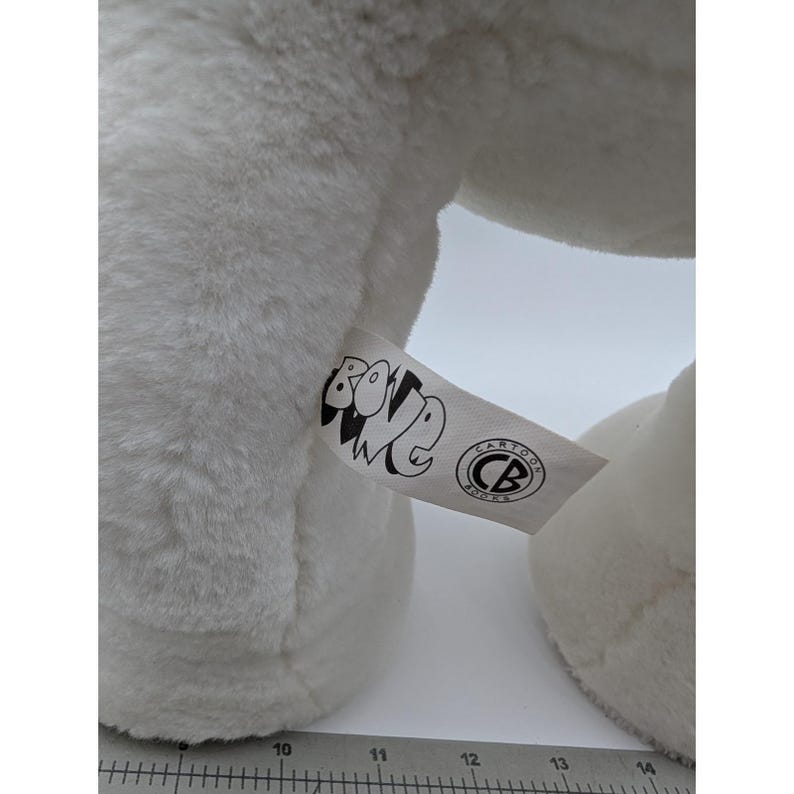 May include: Close-up of a white plush toy with a tag that reads "BONE" and "CARTOON BOOKS" in black text. The toy has a soft, fluffy texture. The tag is white with black text and a circular logo.
