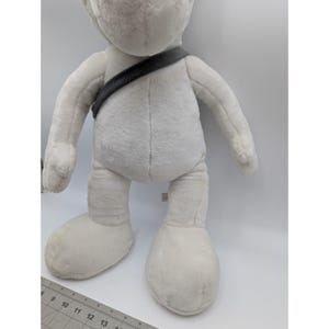 May include: A large, plush, white stuffed animal with oversized feet and a dark gray strap across its chest. The toy has a soft, textured appearance and is standing upright. The background is plain white.