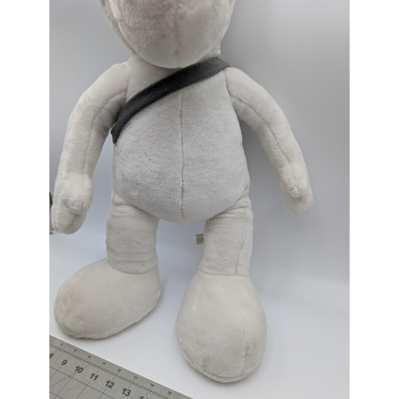May include: A large, plush, white stuffed animal with oversized feet and a dark gray strap across its chest. The toy has a soft, textured appearance and is standing upright. The background is plain white.