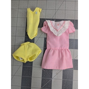 May include: A set of three doll clothes, including a pink gingham dress with a white lace collar, a yellow sleeveless leotard, and yellow shorts.
