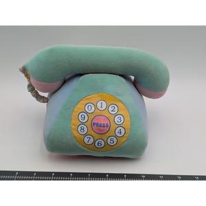 May include: A plush toy telephone in shades of blue, green, and pink. The rotary dial features numbers 0-9 and the word "PRESS" in the center. The handset is curved and attached to the base with a coiled cord.