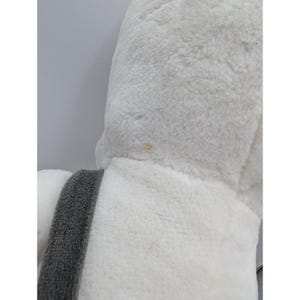 May include: Close-up of a plush, white stuffed animal with a gray accent. The soft toy has a fluffy texture and a small stain on the surface. The gray accent is a strip of fabric.