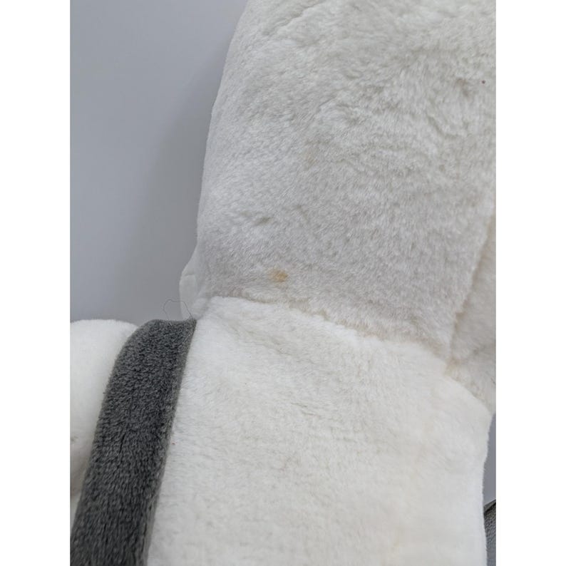 May include: Close-up of a plush, white stuffed animal with a gray accent. The soft toy has a fluffy texture and a small stain on the surface. The gray accent is a strip of fabric.