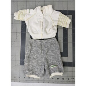 May include: A vintage doll outfit featuring a white jacket with yellow striped sleeves and a gray shorts set. The jacket has a collar and a button closure. The shorts have white trim and a small green label.
