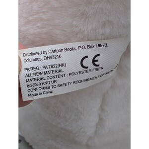 May include: A white tag with black text, attached to a plush toy. The tag includes the distributor's address, material content, and safety information. The tag reads "Distributed by Cartoon Books, P.O. Box 16973, Columbus, OH43216".