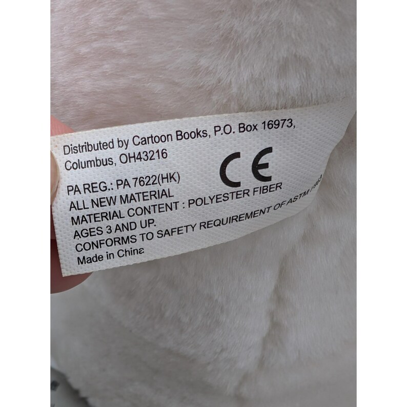 May include: A white tag with black text, attached to a plush toy. The tag includes the distributor's address, material content, and safety information. The tag reads "Distributed by Cartoon Books, P.O. Box 16973, Columbus, OH43216".