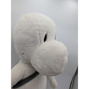 May include: A plush, white cartoon character with a large, rounded snout and black eyes. The stuffed animal has a gray strap across its chest. The toy is made of soft, fuzzy material.
