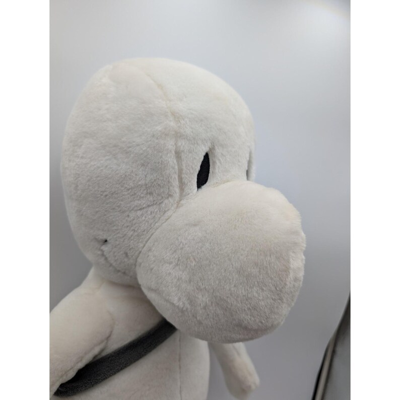 May include: A plush, white cartoon character with a large, rounded snout and black eyes. The stuffed animal has a gray strap across its chest. The toy is made of soft, fuzzy material.