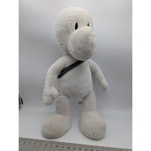 May include: A large, plush, off-white cartoon character doll with a rounded head, large nose, and black eyes. It has a black strap across its chest and large, rounded feet. The doll is approximately 18 inches tall.