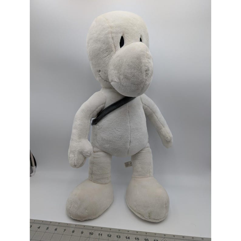 May include: A large, plush, off-white cartoon character doll with a rounded head, large nose, and black eyes. It has a black strap across its chest and large, rounded feet. The doll is approximately 18 inches tall.
