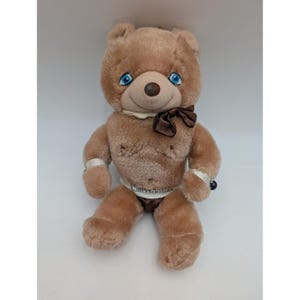 May include: A vintage plush teddy bear with light brown fur, blue eyes, and a brown bow tie. The bear has a white collar and wristbands, and a white diaper with text. A black bead is on the right wrist.