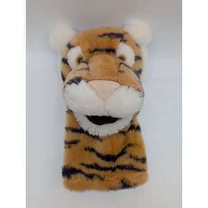 May include: A plush tiger head golf club cover. The cover is primarily tan with black stripes, a pink nose, and white accents around the eyes and ears. The tiger has a smiling expression.