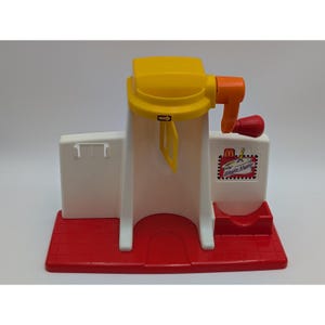 May include: A vintage Shake Maker toy in red, white, yellow, and orange. The toy has a red base, a white structure with a yellow top, and an orange handle with a red crank. The Shake Maker logo is visible on the side.
