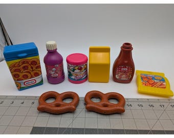 May include: A collection of colorful toy food items, including a blue box of cookies, a purple bottle, a pink can, a yellow milk carton, a brown bottle, a yellow bag of chips, and two brown pretzels. The toys are arranged on a white surface.