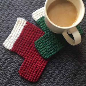 May include: Two knitted Christmas stocking-shaped coasters, one red and white, one green and white, with a white mug filled with coffee on top of the green coaster.
