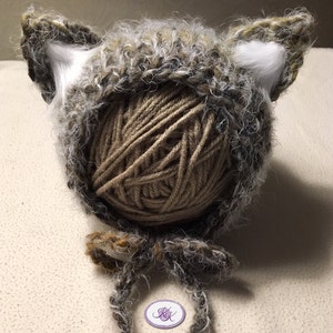 May include: A brown and gray crocheted baby hat with white fur ear flaps. The hat is made with a fuzzy yarn and has a ball of yarn in the center.