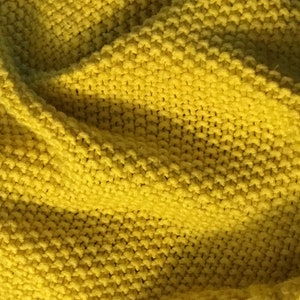 May include: Close-up of a soft, yellow knitted fabric. The texture is visible, with a repeating pattern of raised stitches. The fabric appears to be thick and warm, suggesting a blanket or scarf.