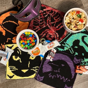 May include: A collection of knitted Halloween-themed squares featuring black cats, bats, and pumpkins. The squares are arranged on a wooden surface with candy, a bowl of M&Ms, and a bag of Reese's Pieces. The squares are all different colors, including black, purple, red, and green.