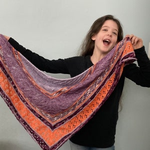 May include: A purple and orange knitted shawl with a lacy pattern. The shawl is draped over the shoulders of a person wearing a black long-sleeved shirt.