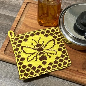 Op de afbeelding: Een geel en bruin gebreide pannenlap met een bijenontwerp. De vierkante pannenlap heeft een ophanglus. Een glazen pot met vloeistof en een roestvrijstalen pot zijn ook zichtbaar.