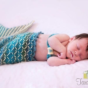 May include: A newborn baby wearing a knitted mermaid tail costume in shades of blue, green, and white, sleeping on a white blanket.