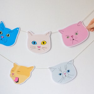 Puede incluir: Una cadena de cinco recortes de caras de gato, cada uno con una expresión diferente. Los gatos son azules, blancos, rosas, naranjas y grises. La cadena es blanca y los recortes están unidos con pequeños agujeros.
