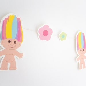 May include: A colorful garland with two rainbow-haired trolls and pink, green, and blue flowers. The garland is perfect for a birthday party or a nursery.