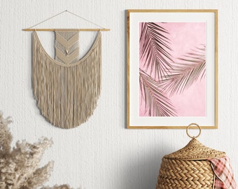 May include: A beige macrame wall hanging with a geometric pattern, a framed print of palm leaves on a pink background, and a woven wicker laundry basket with a plaid throw draped over the top.