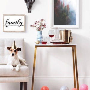 May include: A white dog wearing a party hat sits on a light beige armchair. A gold metal table with a white marble top is in the background. There are two wine glasses, a decanter, and a gold ice bucket on the table. There are also two framed pictures on the wall behind the table. One picture is a black and white image of a bull skull. The other picture is a landscape photograph of a forest. The text "family where life begins & love never ends" is printed on a white frame.