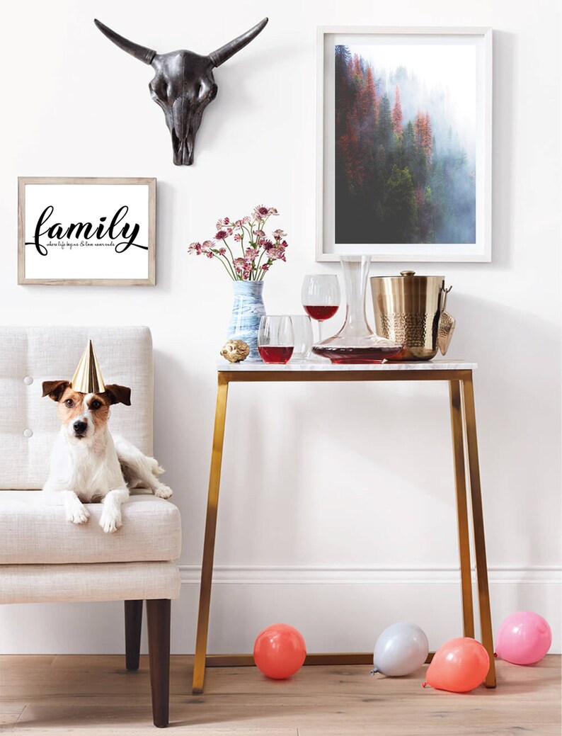 May include: A white dog wearing a party hat sits on a light beige armchair. A gold metal table with a white marble top is in the background. There are two wine glasses, a decanter, and a gold ice bucket on the table. There are also two framed pictures on the wall behind the table. One picture is a black and white image of a bull skull. The other picture is a landscape photograph of a forest. The text "family where life begins & love never ends" is printed on a white frame.