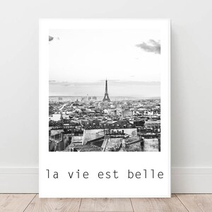 May include: Black and white photograph of the Eiffel Tower in Paris, France. The photo shows the tower from a distance, with the city skyline in the background. The text "la vie est belle" is printed below the image.