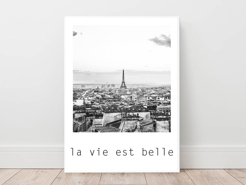 May include: Black and white photograph of the Eiffel Tower in Paris, France. The photo shows the tower from a distance, with the city skyline in the background. The text "la vie est belle" is printed below the image.