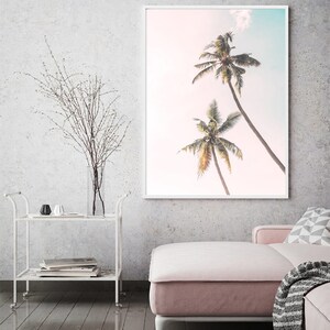 May include: A framed print of two palm trees against a pink and blue sky. The print is hanging on a white wall in a living room setting.