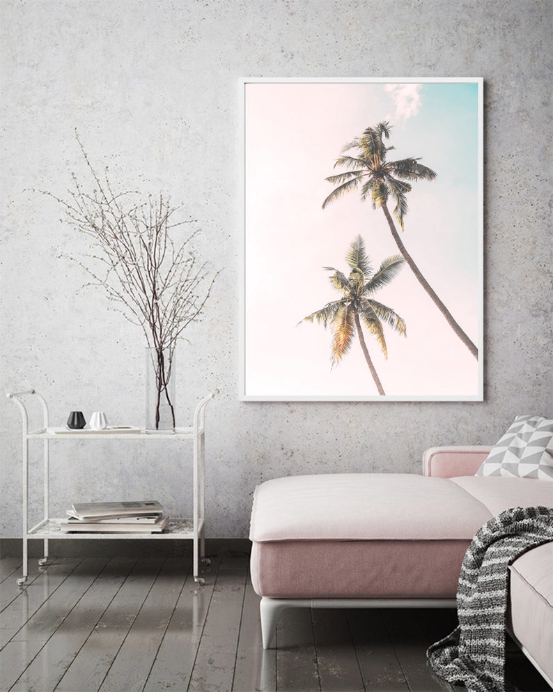 May include: A framed print of two palm trees against a pink and blue sky. The print is hanging on a white wall in a living room setting.