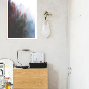 May include: A framed photograph of a misty forest with red and green trees. The photo is hanging on a white wall above a wooden dresser with three drawers. A black lamp and a magazine are on top of the dresser.